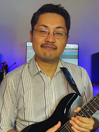 Portrait of Chris Takita, a musician, holding an acoustic guitar and smiling towards the camera with a blurred background.